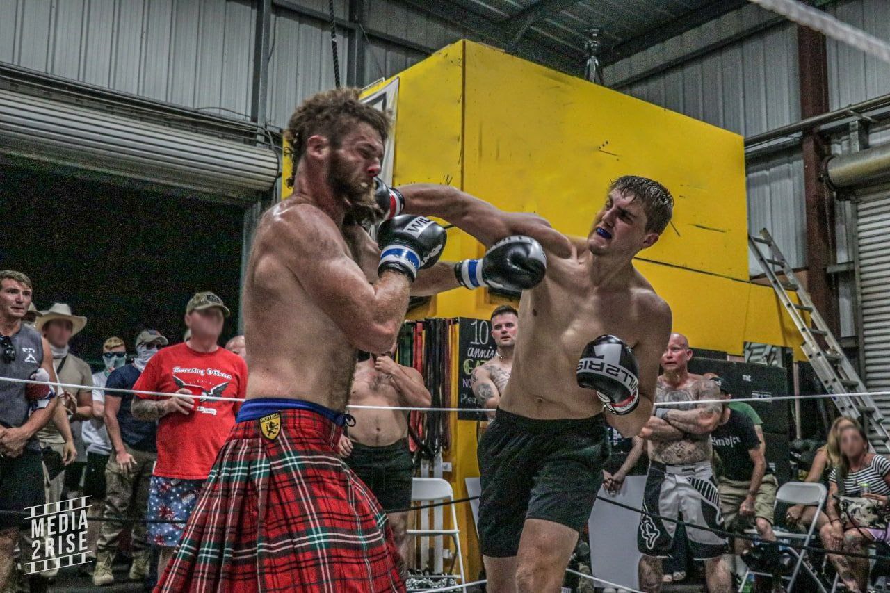 Two sweaty and shirtless men wearing matching black and white Will2Rise branded boxing gloves compete in a one-on-one fight. A small group of white men surround one side of the makeshift boxing ring and look on in excitement. At the exact moment this photograph was taken, the fighter on the right (wearing black shorts) has connected his right fist to the jaw of his opponent, a muscular fellow who dons a red tartan kilt with a hint of blue undershorts peeking from below the waistband. Two female spectators are watching from the folding chairs while some of the other would-be fighters wait for their chance to get knocked out.