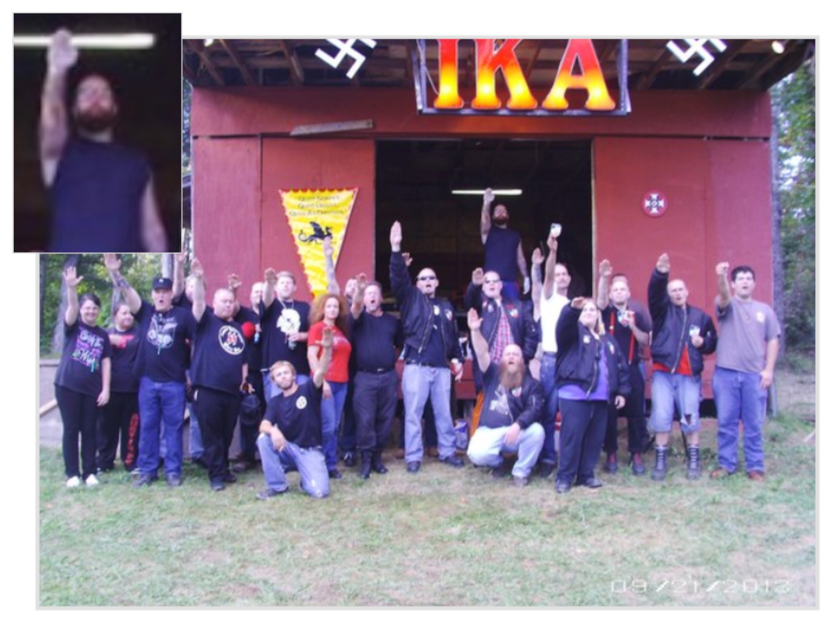 A large group of people, arms raised in a “Sieg Heil” salute, are assembled in front of a small, haphazard appearing red building. An illuminated sign bearing the letters “I.K.A.”, centered between two smaller white swastikas, hangs over the group. At the back of this group, a man wearing a navy tank top is seen standing on an unidentified object, appearing above the others. 