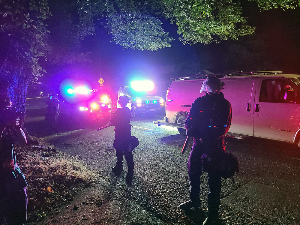 Officers stand, highlighted by cherry lights in the darkness, ready to brutalize the citizenry of Portland that is rising up against their brutality.