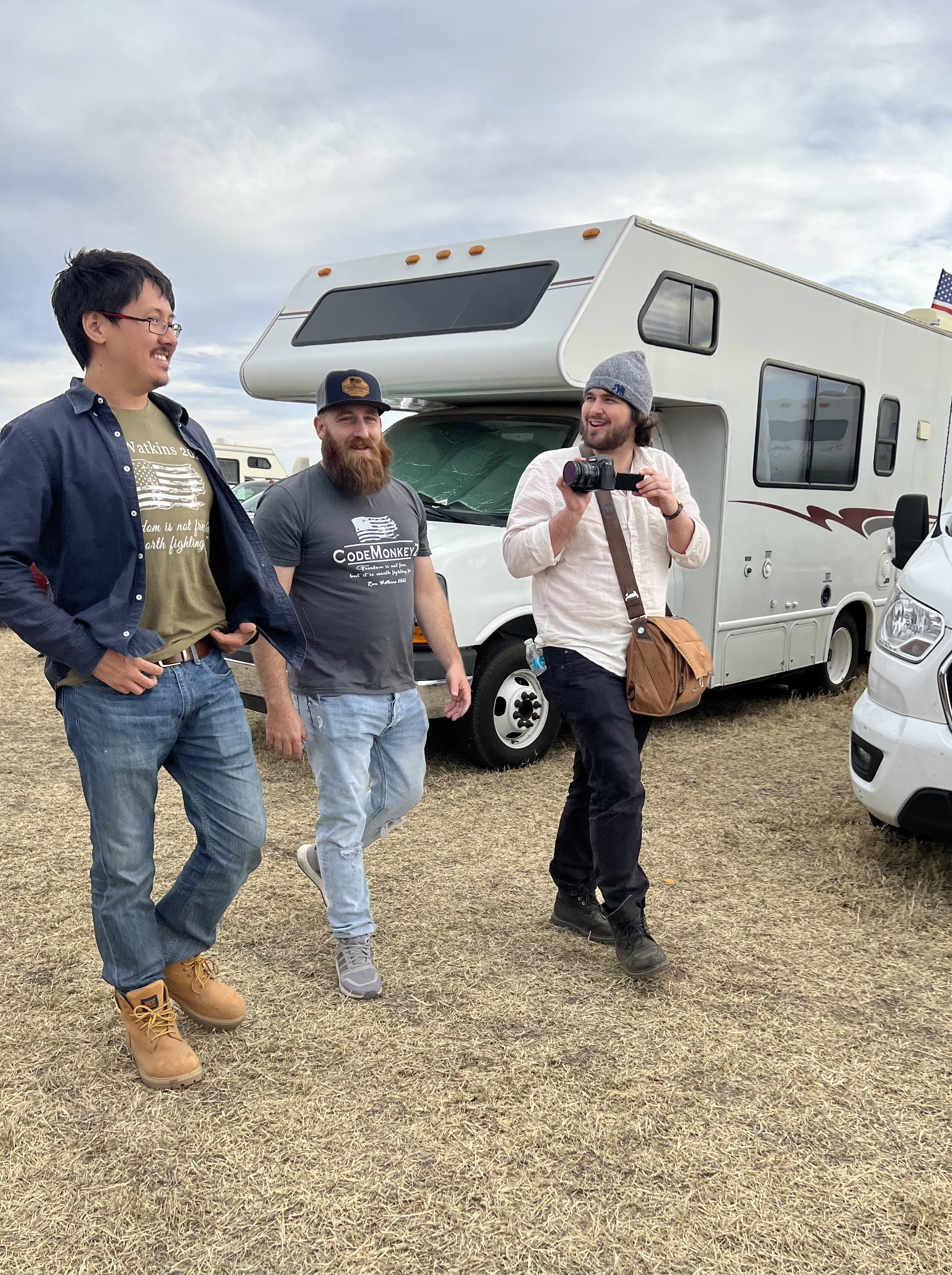 A smiling Ron Watkins on the left walks and talks with two other men as they pass trailers and booths in the desert