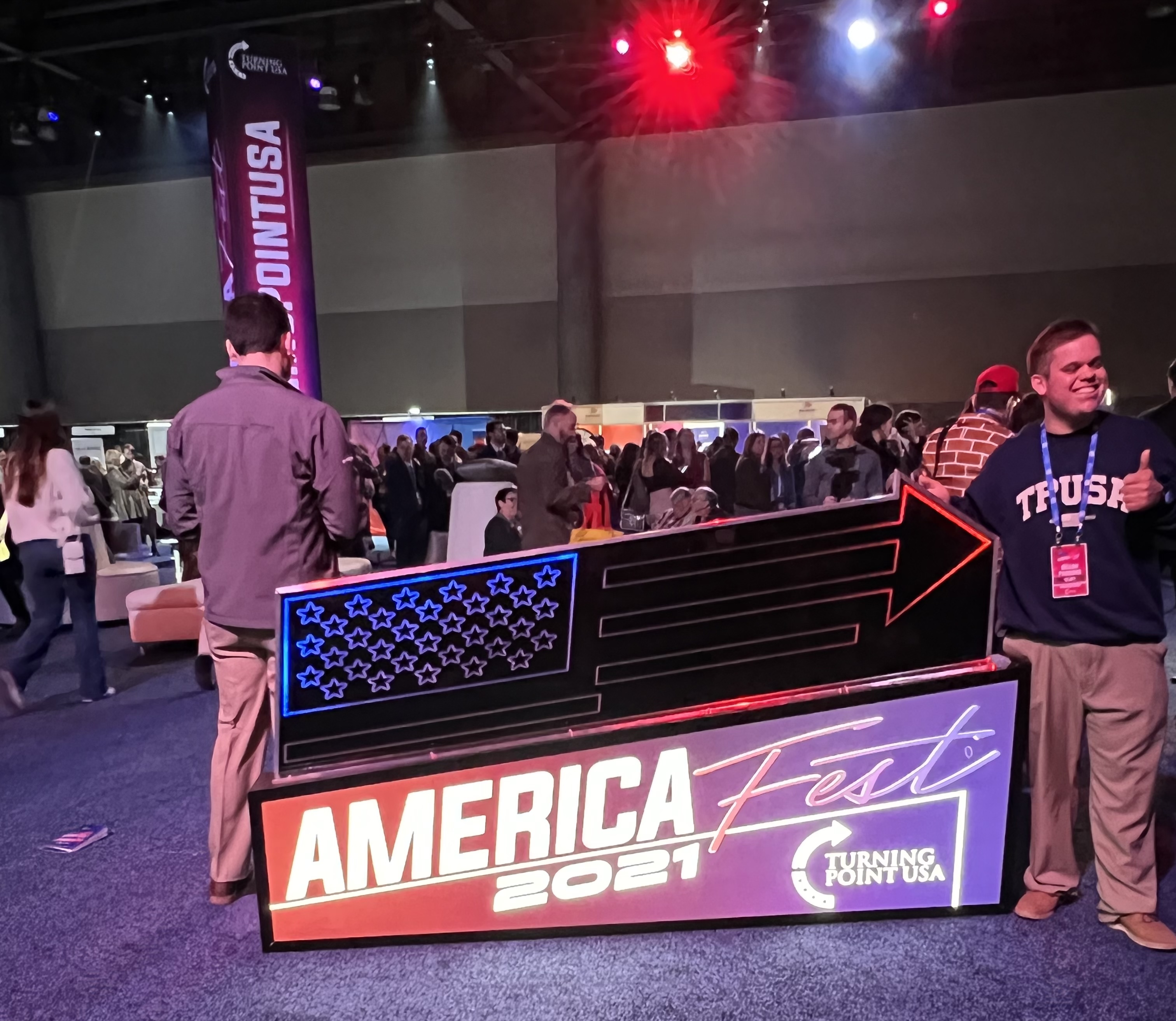 A young man in a TPUSA sweater poses next to a neon AmericaFest 2021 sign in the exhibitor hall. A crowded group of people walk around the hall in the background, including a man in a brick pattern suit. The room is lit up with red, white, and blue