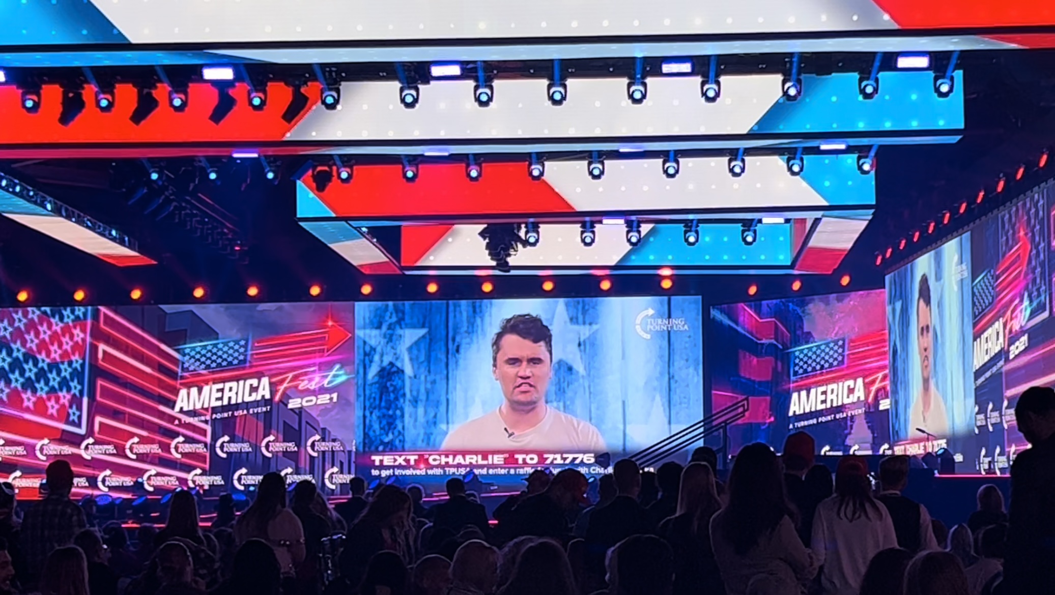 Two large video screens show Charlie Kirk advertising a lunch date with him if you 'text CHARLIE to 71766 to get involved with TPUSA and enter a raffle to have lunch with Charlie Kirk.' The stage lights are red, white and blue, the backdrop has advertisements for AmericaFest and TPUSA