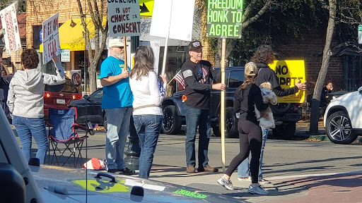 mostly white people waving american flags and holding up antivaxx signs on a street corner