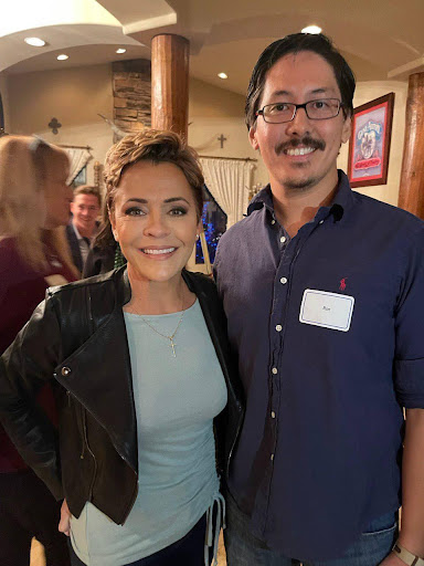 2022 gubernatorial candidate Kari Lake smiles for a photo with Ron Watkins, who is also smiling. The both stand in a cross-heavy room for one of her campaign dinners