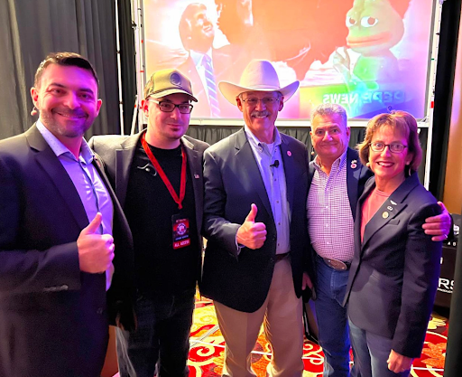 Arizona republicans pose for a photo with QAnon John Sabal. In order: Rep. Leo Biascuicci, QAnon John, Rep. Mark Finchem, Sen. Sonny Borrelli, and Sen. Wendy Rogers. Behind them a video plays with Trump and Pepe on the screen