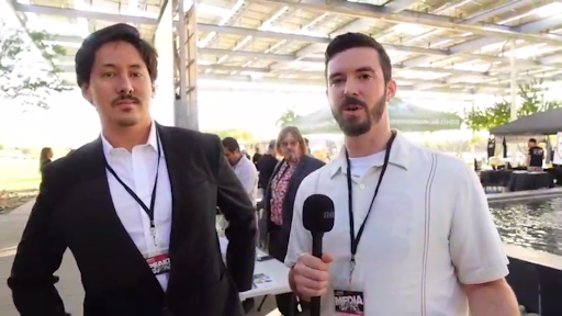 Ron Watkins stands in a suit looking into the camera, as he gives an interview with Greyson Arnold who’s holding a microphone and has a media pass around his neck. Jim Watkins stands behind them staring