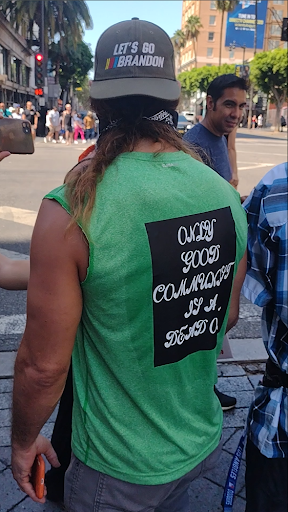 A man in a green sleeveless shirt stands with his back to the camera. The back of his shirt reads “The only good communist is a dead one” and he is wearing a hat that says “Let’s Go Brandon.