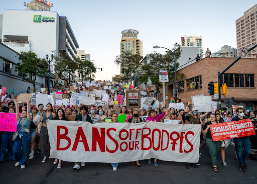 Thousands of protesters march through downtown San Diego at dusk, occupying the breadth of the street. A banner is held by six people at the front of the crowd which reads, “Bans Off Our Bodies.” 
