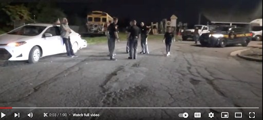 Several police officers are standing in a parking lot with members of the people’s convoy. A police cruiser is visible to the left, assorted convoy vehicles including a schoolbus are also in view.