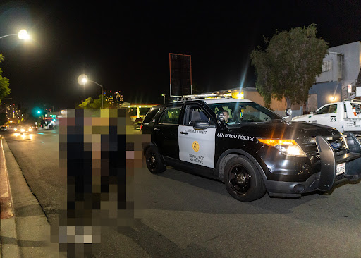 two protesters, pixelated to protect their identity, engage with an SDPD cruiser which has stopped in the street. Officer Devor is at the wheel.