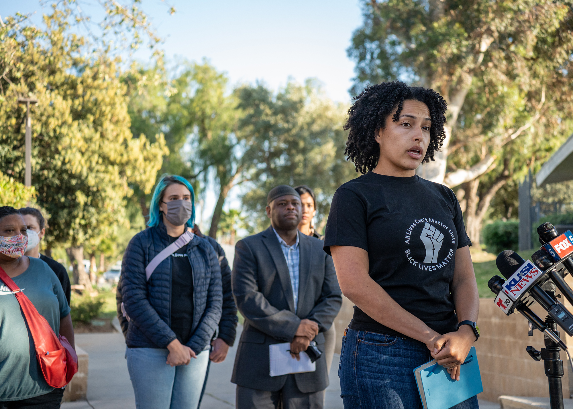 Danielle Wilkerson, a young black woman speaks to reporters in a shirt that reads 'all lives can’t matter until black lives matter.' It is early evening