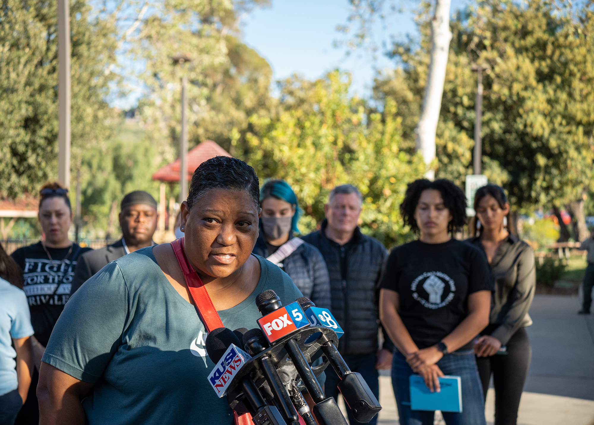 Tasha Williamson stands before a crowd in the evening sun to speak to reporters who are off-screen. In the crowd, an activist holds a sign that says 'Protect Black Women.'