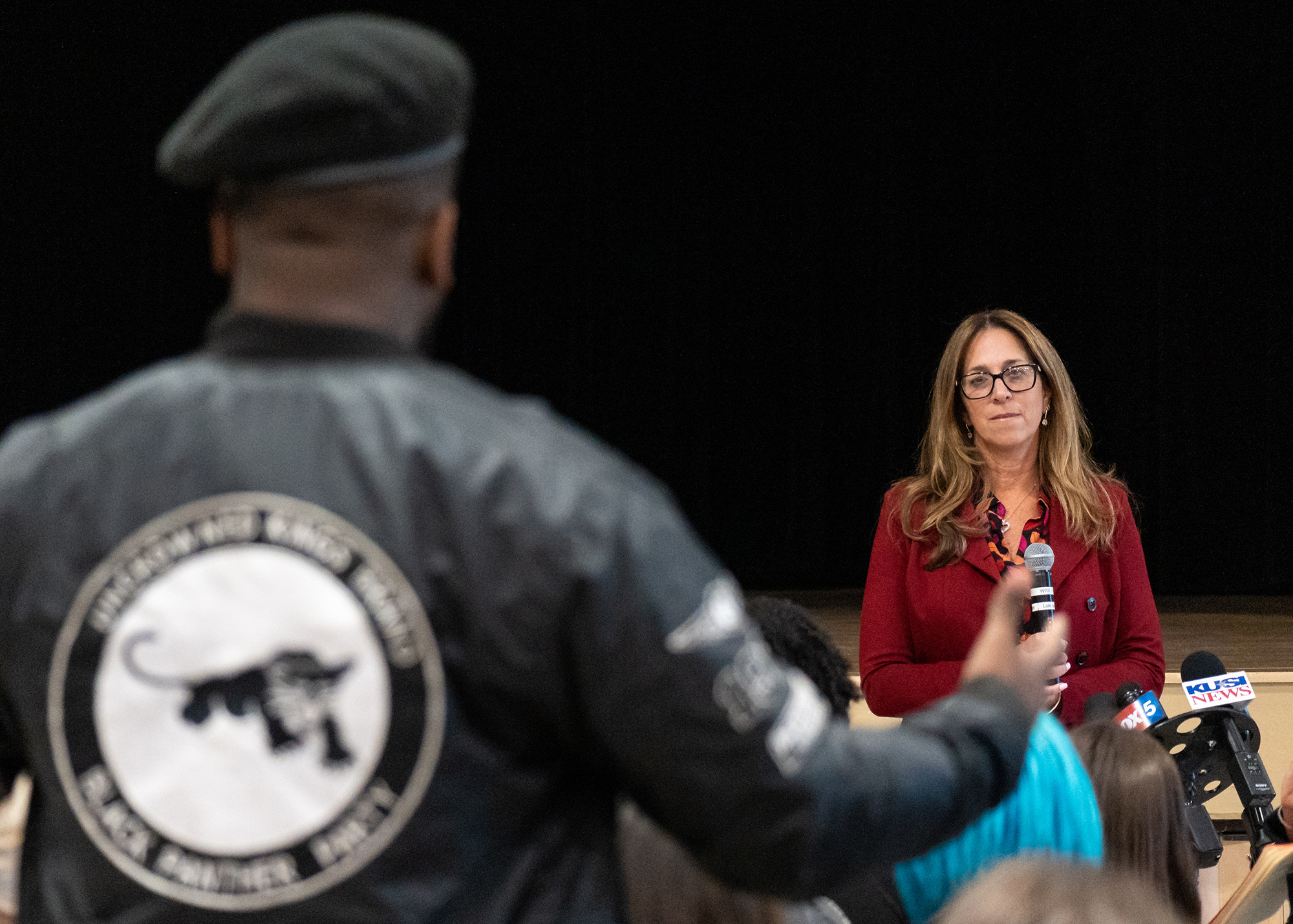 Lisa Weinreb wears a red jacket while holding a microphone. She is listening to a member of the Black Panther Party of San Diego, who is asking a question and gesticulating in the foreground. He is out of focus, and his hand overlaps with the distant microphone.