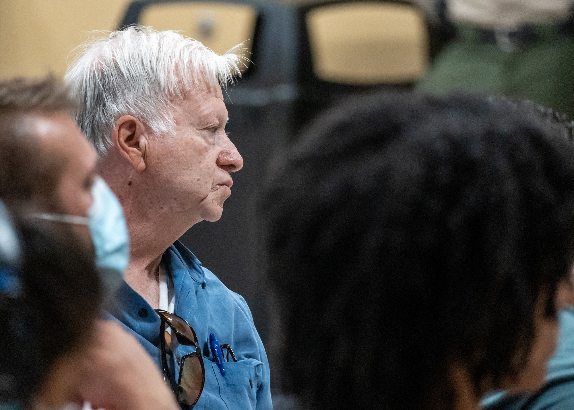 An elderly man looks to the right of the frame, in profile, as numerous figures are out of focus in the foreground.