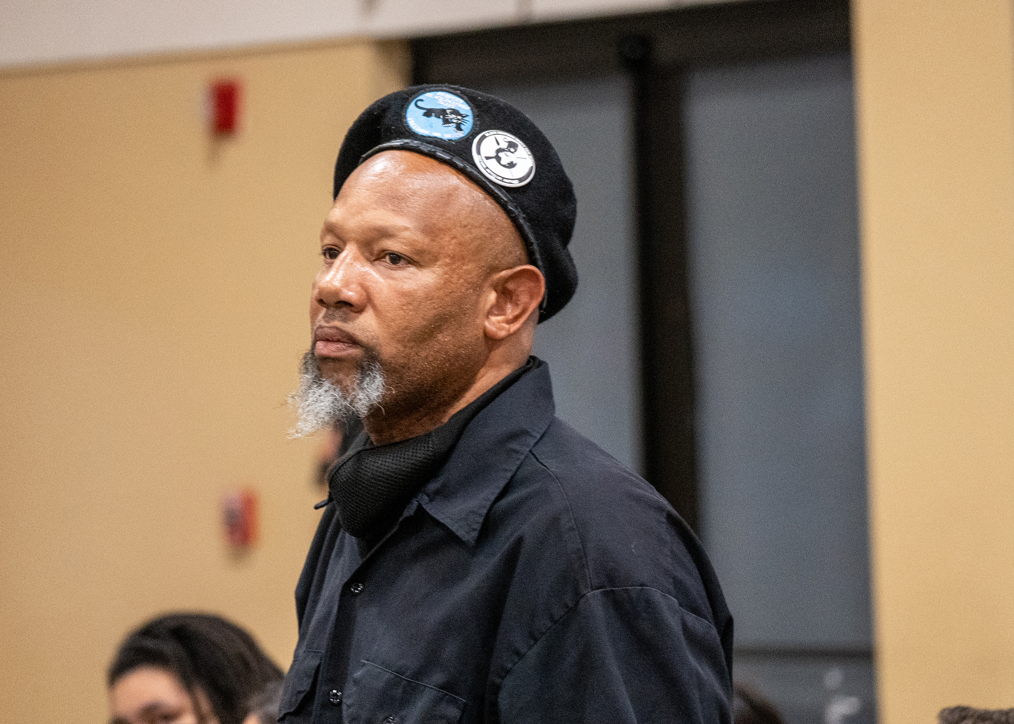 A man with a graying beard, a black beret with Black Panther pins, and a black shirt glares at Lt. Wray, who is out of frame. The man is a member of the Black Panther Party of San Diego.