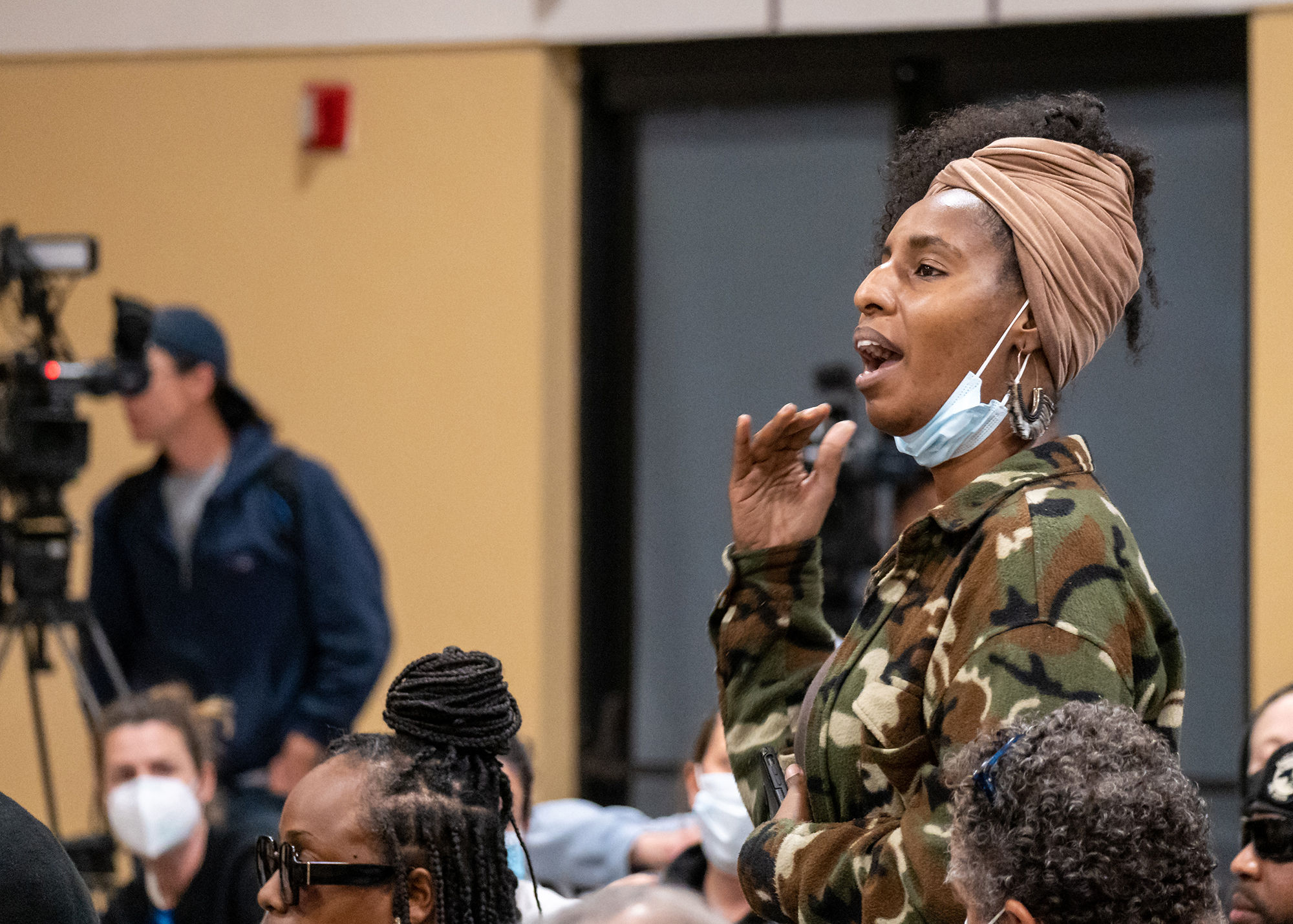 an attendee in a camouflage jacket stands to address Lt. Wray while gesturing. A television camera is out of focus in the background, behind the crowd.