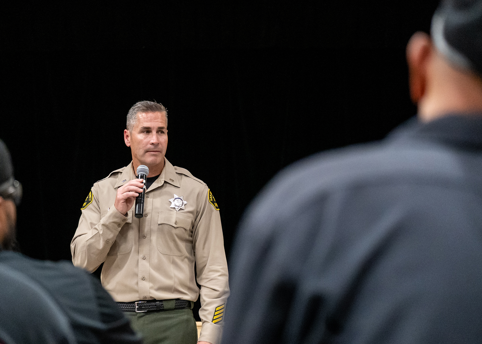 Lt. Wray is framed between two sitting figures in the foreground. Behind Wray is the black curtain of the gymnasium’s stage.