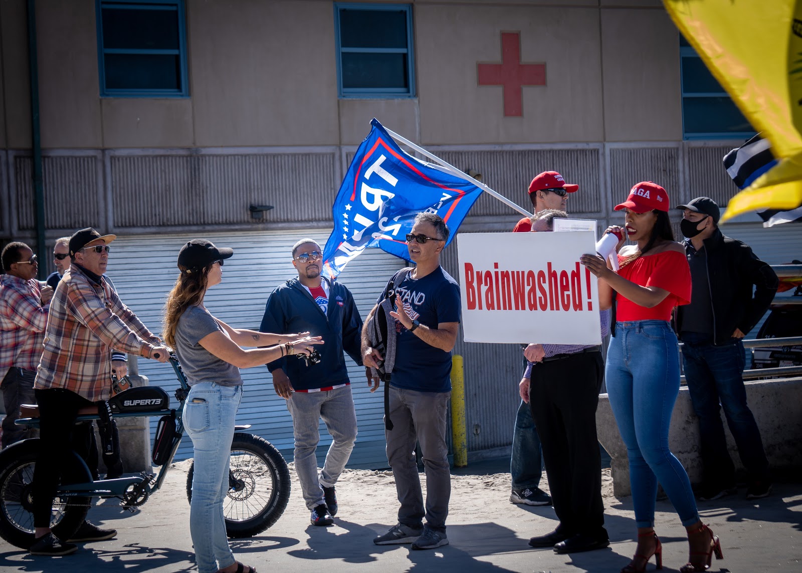 A passerby confronts the demonstrators--primarily the individual in the maga hat with the Brainwashed sign–and Joe Collins is visible in full observing the discussion. Vignette added for emphasis.