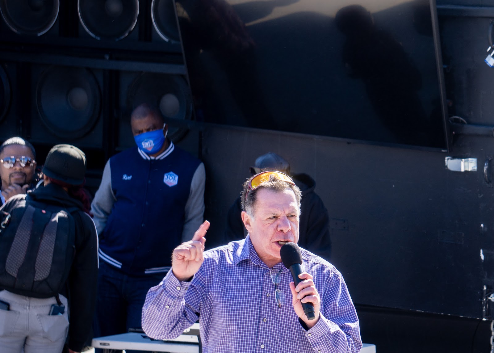 The previously mentioned unidentified speaker is talking into a microphone. Joe Collins appears, incidentally, at the left edge of the frame, with a photographer and someone on his staff complete with matching Joe Collins jackets.