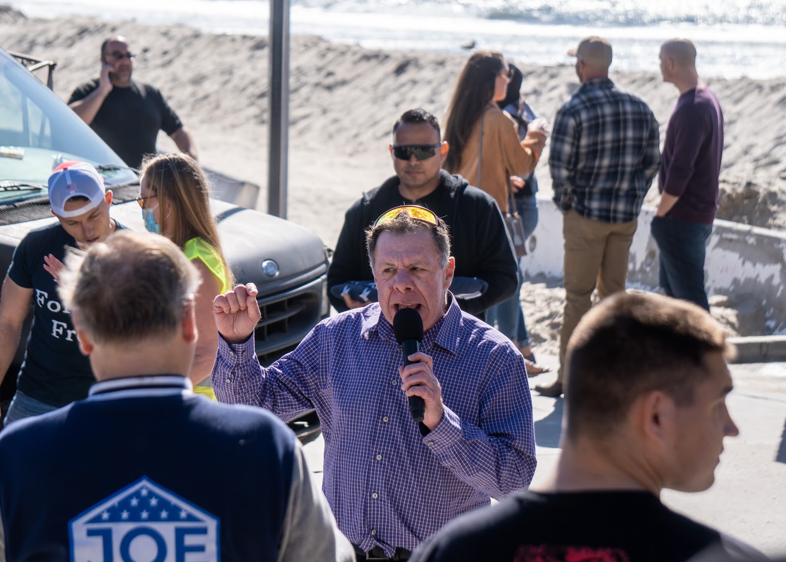 An unidentified speaker nearly yells into a microphone while holding up a fist to gesticulate and emphasize his points. In the foreground is a young man and an individual in a Joe Collins jacket, out of focus.