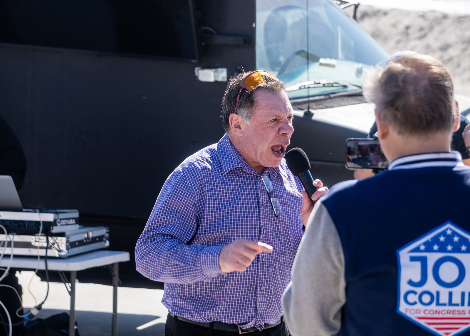 The same unidentified speaker yells into a microphone and points his finger for emphasis while the same Joe Collins employee is visible in the foreground.