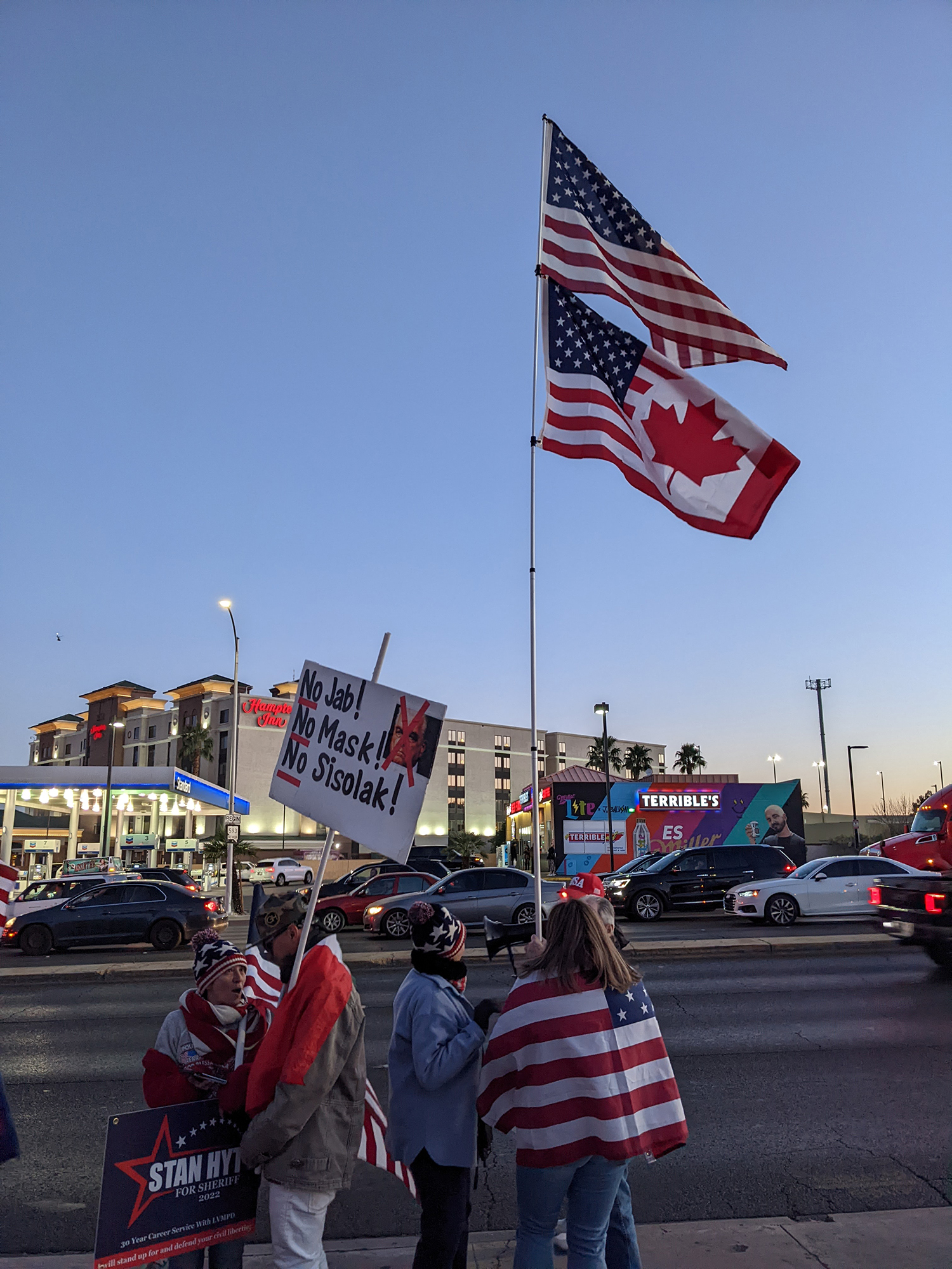 people holding a flag pole with a hybrid canadian american flag on it and another person with a sign that says no jab! no mask! no Sisolak!