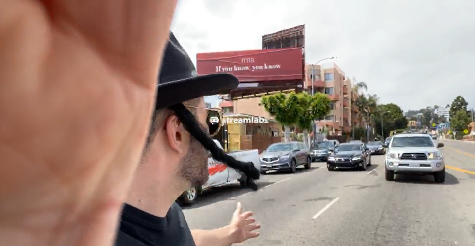 a man wearing a mocking imitation of a hasidic jewish man's dress holds his hand on a camera and looks out at traffic as he stands in the street