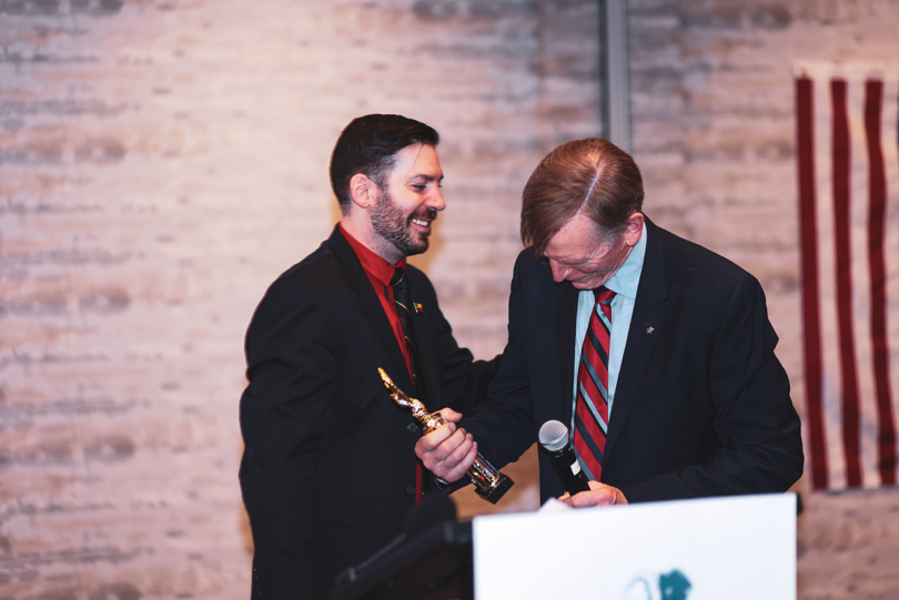 Republicans for National Renewal leader, Mark Ivanyo, gives Rep. Paul Gosar the made-up 'America First' award. Gosar looks down at the award smiling as Ivanyo pats his back.