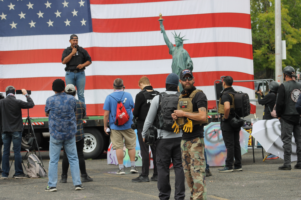 A tall man stands with a microphone on a stage with a giant american flag behind him with a person-sized statue of liberty model off to the side while men in body armor mill about