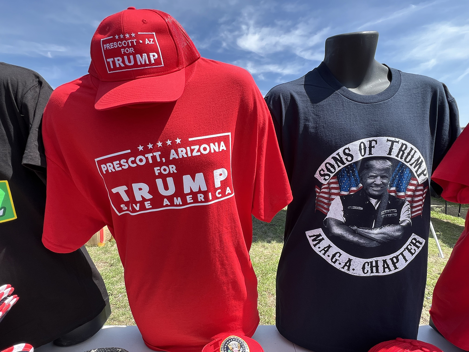 two shirts and a hat on sale being displayed at the merch booths. One shirt is bright red with white text reading 'Prescott, Arizona for Trump. Save America.' The red hat sitting on top matches the shirt in color and text. Another shirt being displayed next to the red shirt is a black spin on a Sons of Anarchy shirt with Trump photoshopped as a biker. The text reads 'Sons of Trump, MAGA Chapter.'