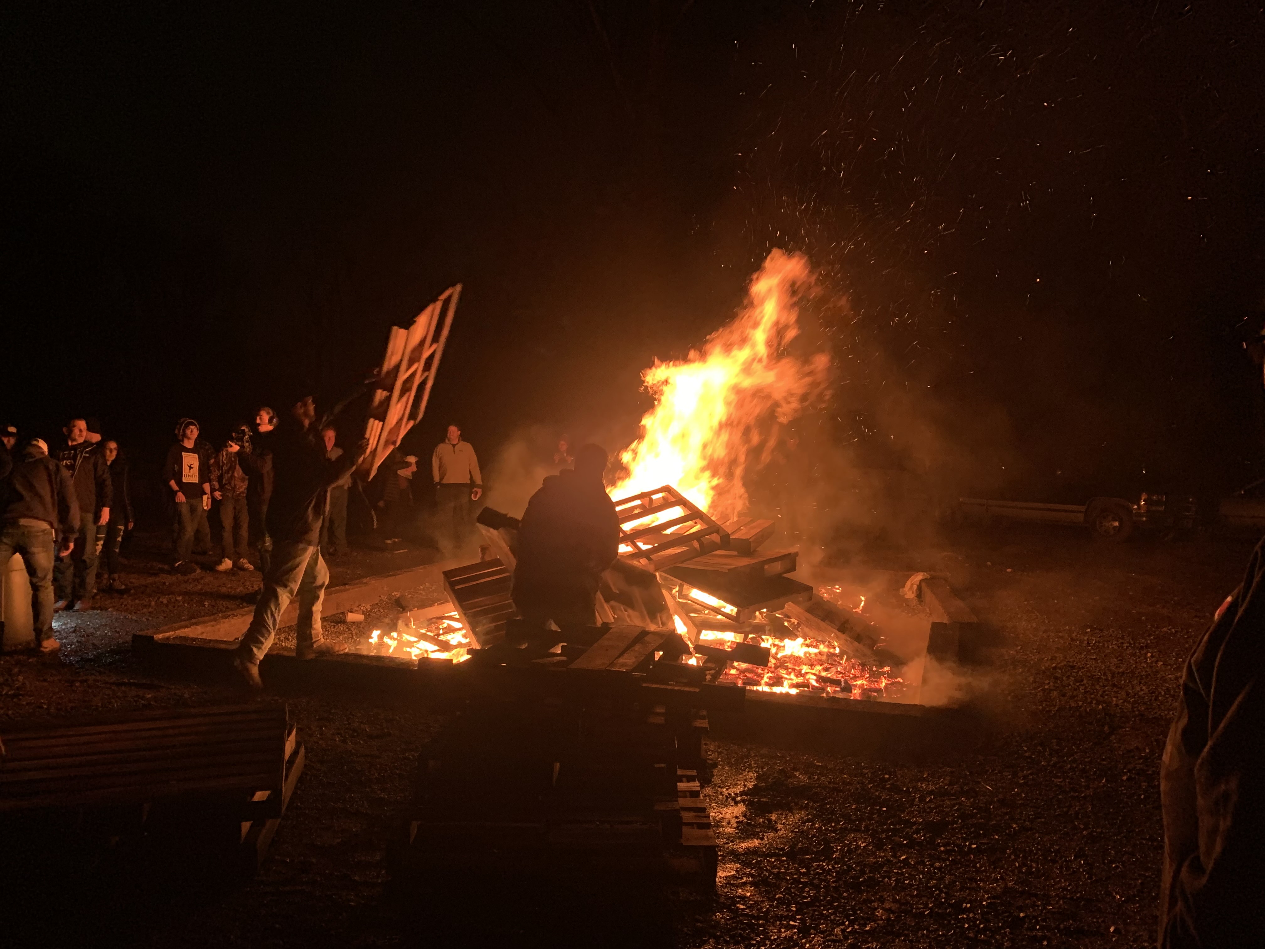 A man throws a palette into a bonfire as many people gather around