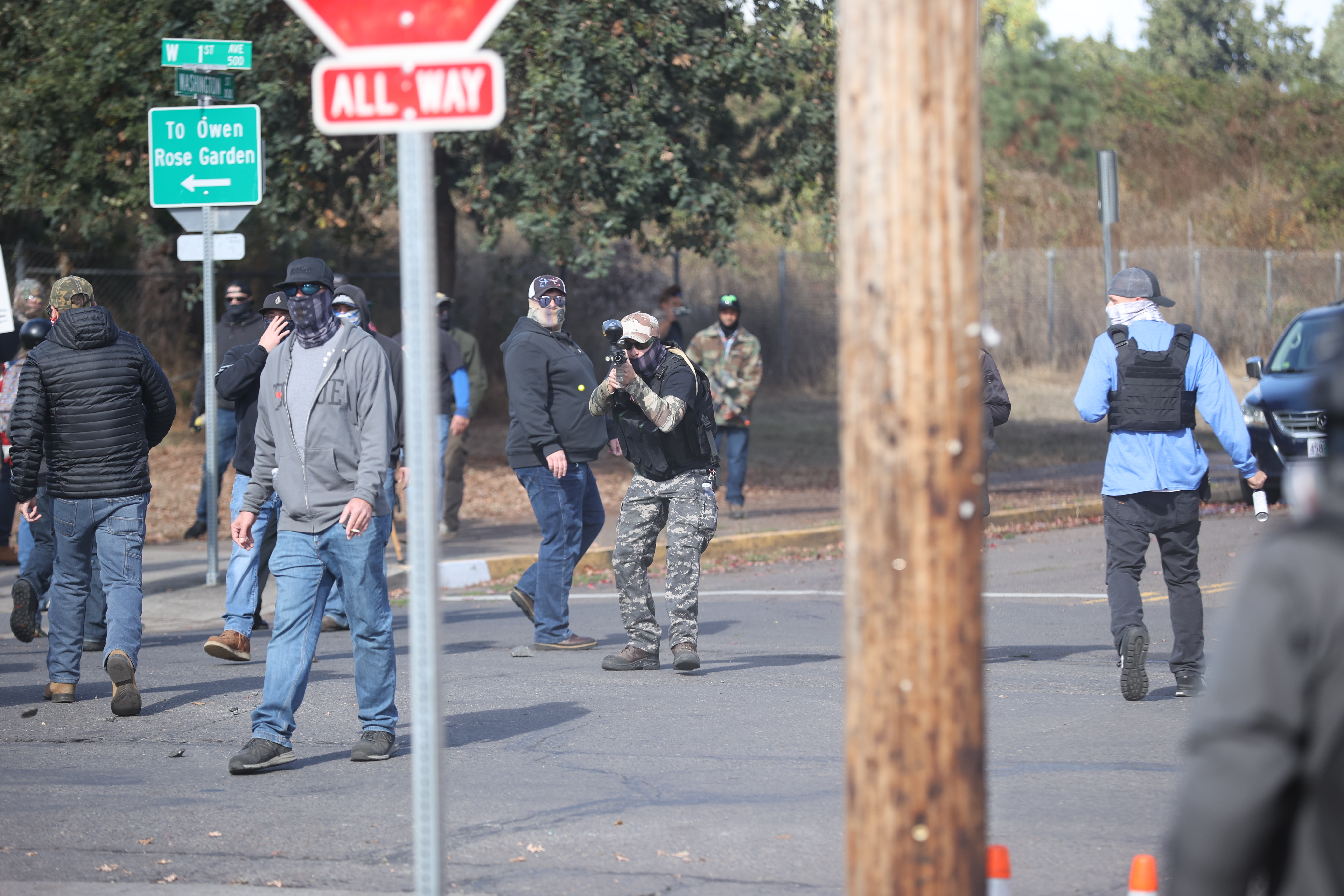 A color photograph looking past a blurry red stop sign and wooden electrical pole. Beyond the signs are men, with their faces covered, retreating on the 1st Ave. To the right of the wooden pole is the blue-shirted Mustin walking away. Directly in the middle of the shot is Jonathan Donovan pointed his paintball gun directly at the camera this photo is taken from. Around him and to the left are another dozen or so protesters leaving.