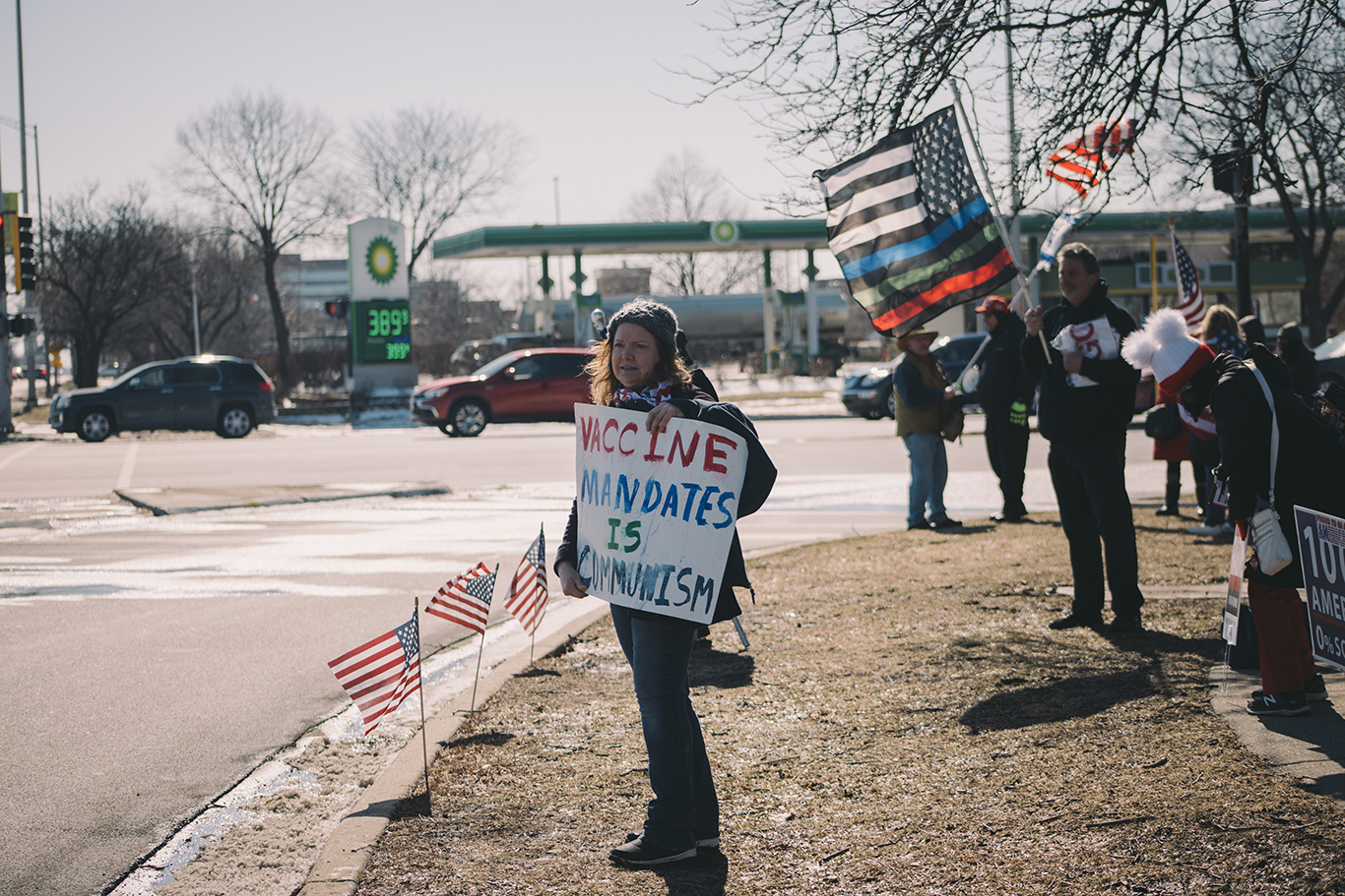 a woman stands with a sign that says 'vaccine mandates is communism' while others behind her wave flags on the side of a road