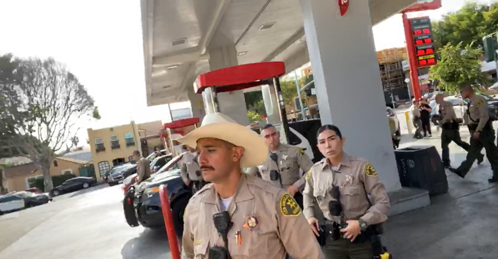 several sheriffs mill about a gas station, one in a cowboy hat in front with a female sheriff's officer behind him