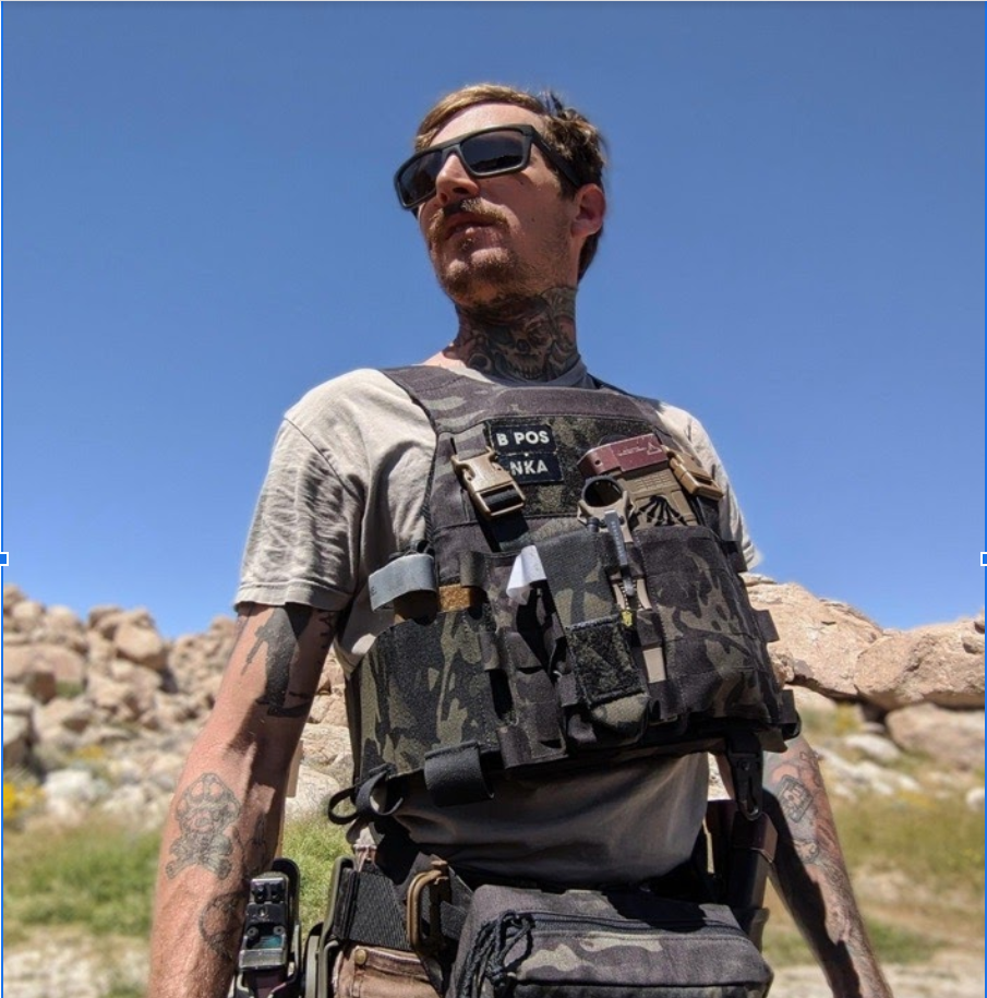 A man poses in the desert with body armor and a handgun in a holster on his leg.