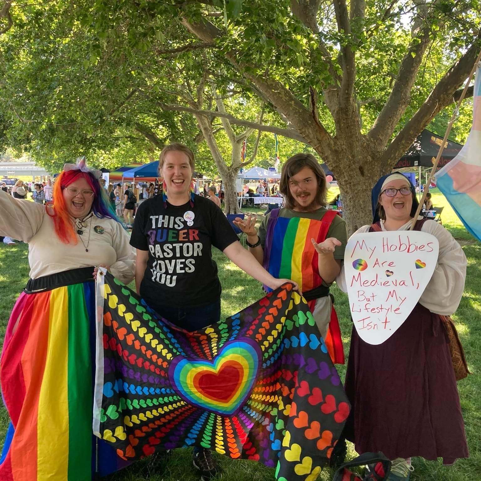some of the attendees in Rennaisance garb with rainbow flags and decor. One wears a shirt that says 'This queer pastor loves you' and another has a sign that says 'my hobbies are medieval but my lifestyle isn't'u