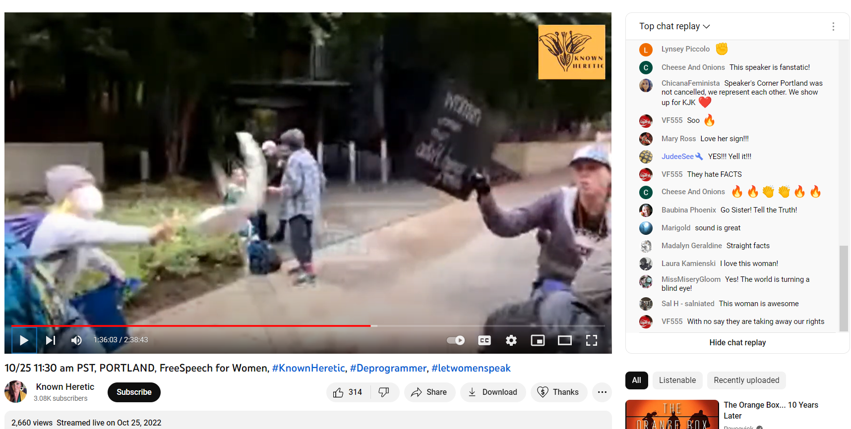 A screen capture of a YouTube video. A cream pie is in the center of frame, being thrown by a person whose identity is disguised. A woman at the right attempts to use her transphobic sign to block