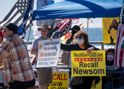 a close up shot of several people, two men are maskless, and in the background there are thin blue line american flags in the background. a masked woman holding up two signs, the one in her right hand reads: 'CNN: Communist News Network! China News Network! MSM: Main stret media! CNN MSM would be in the toilet without Donald J Trump!!!' in bold text on a white backround the other sign reads: 'RecallGavin2020.com, Recall Newsom' it is in bold black text on a rellow backround. there is a red star design dividing the two texts from each other