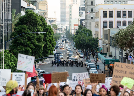 The foreground of the image is the top of a crowd of protesters, out of focus. In focus, though far from the camera, is a line of bike cops standing with police cruisers behind them.