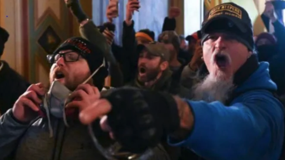 two men shout at the front of a crod inside the capitol building, one on the right in a blue hoodie and oath keepers hat pointing