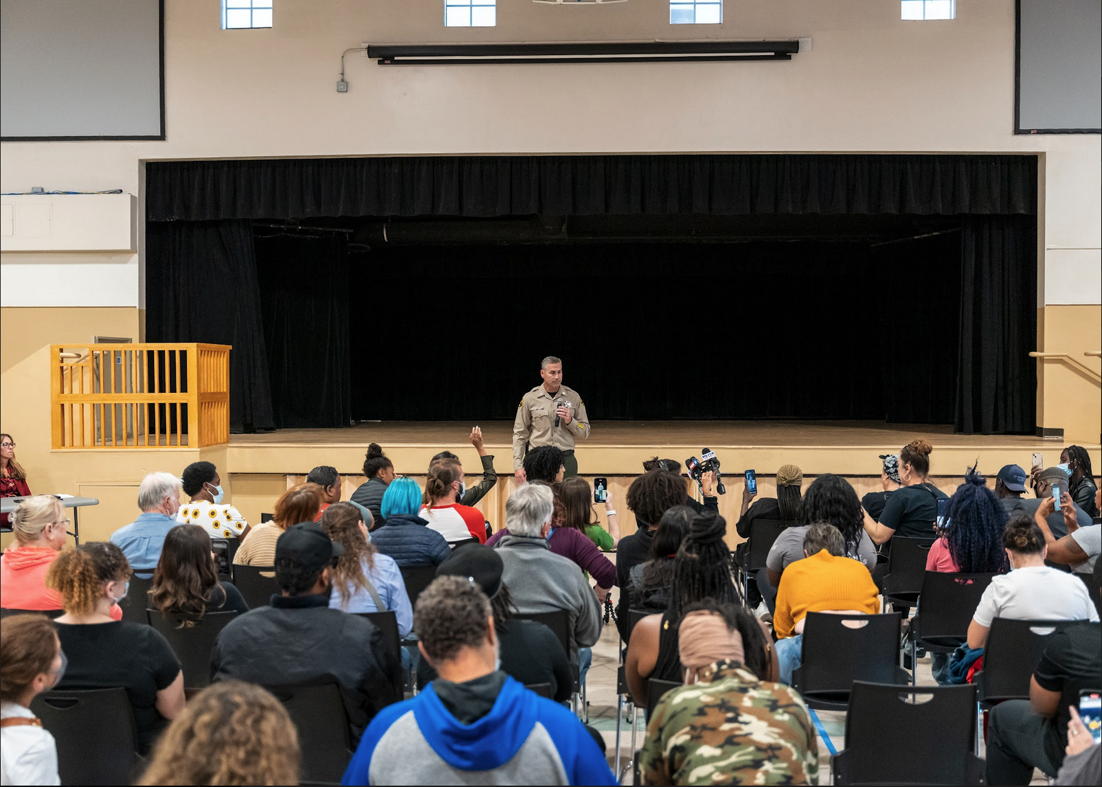 A crowd of sitting people extends beyond the left, right, and bottom sides of the frame. Lt. Wray is visible standing at the far end of the crowd, addressing the gymnasium.