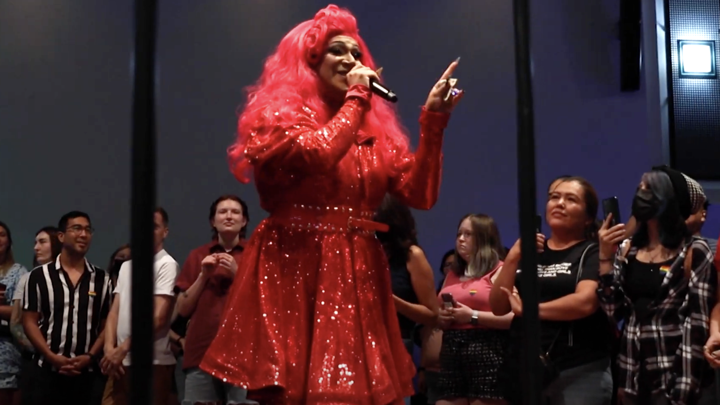 drag queen Pyraddiction is dressed in a bright red wig and bright glittery red long-sleeve dress as she smiles and holds a mic while addressing the crowd. The crowd looks on, many holding up with phones filming, and listens to Pyraddiction speak.