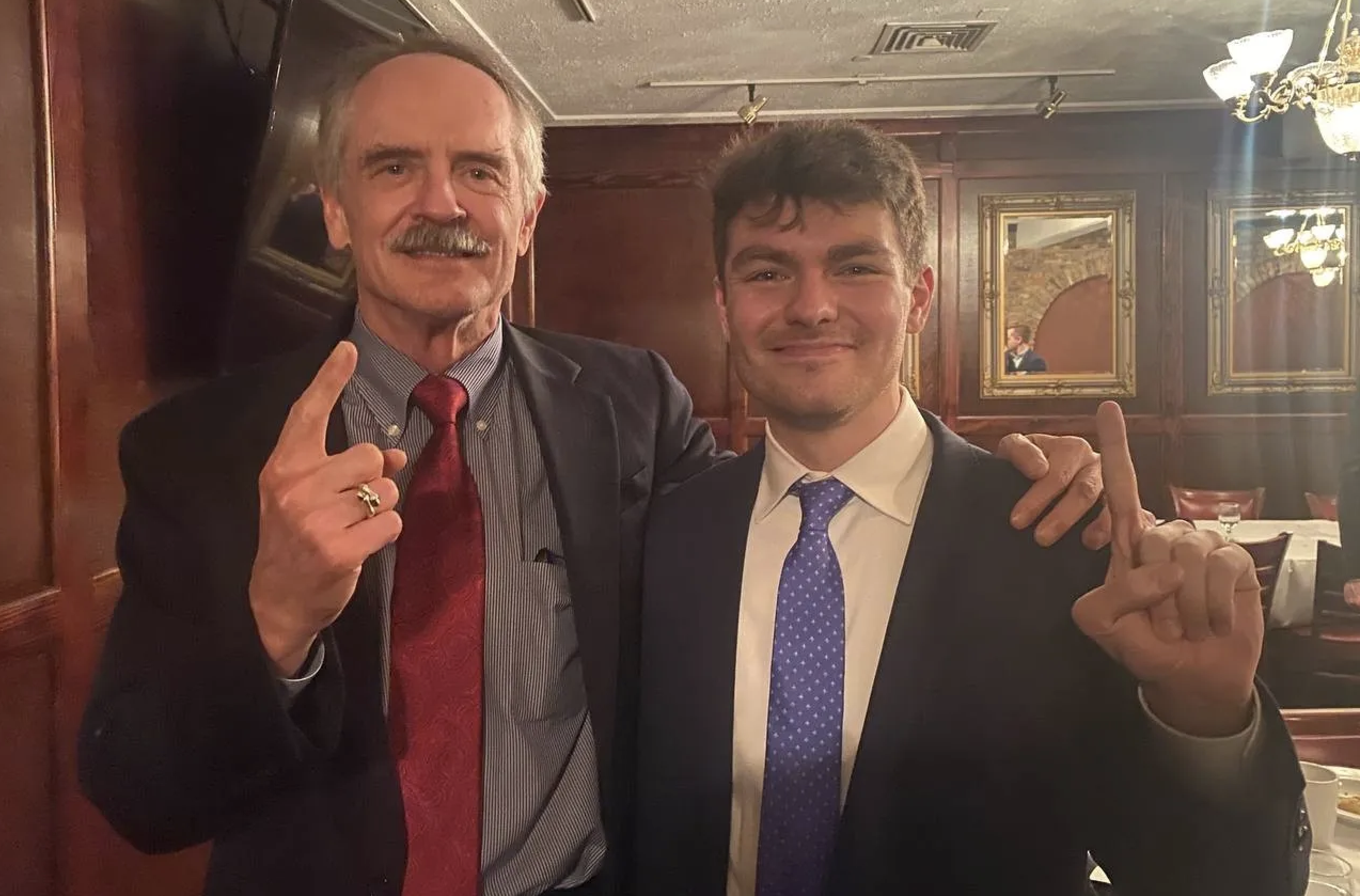 Jared Taylor and Nicholas Fuentes posing for a photo in a room with hardwood walls and a chandelier, both men are in a suit and tie and holding up their index finger pointing up.