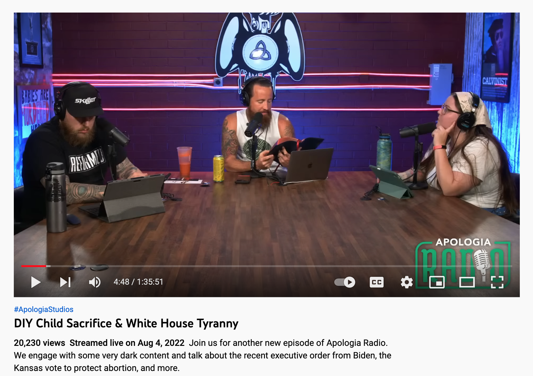 two bearded men and a woman sit at a blue-litten desk with microphones in front of them