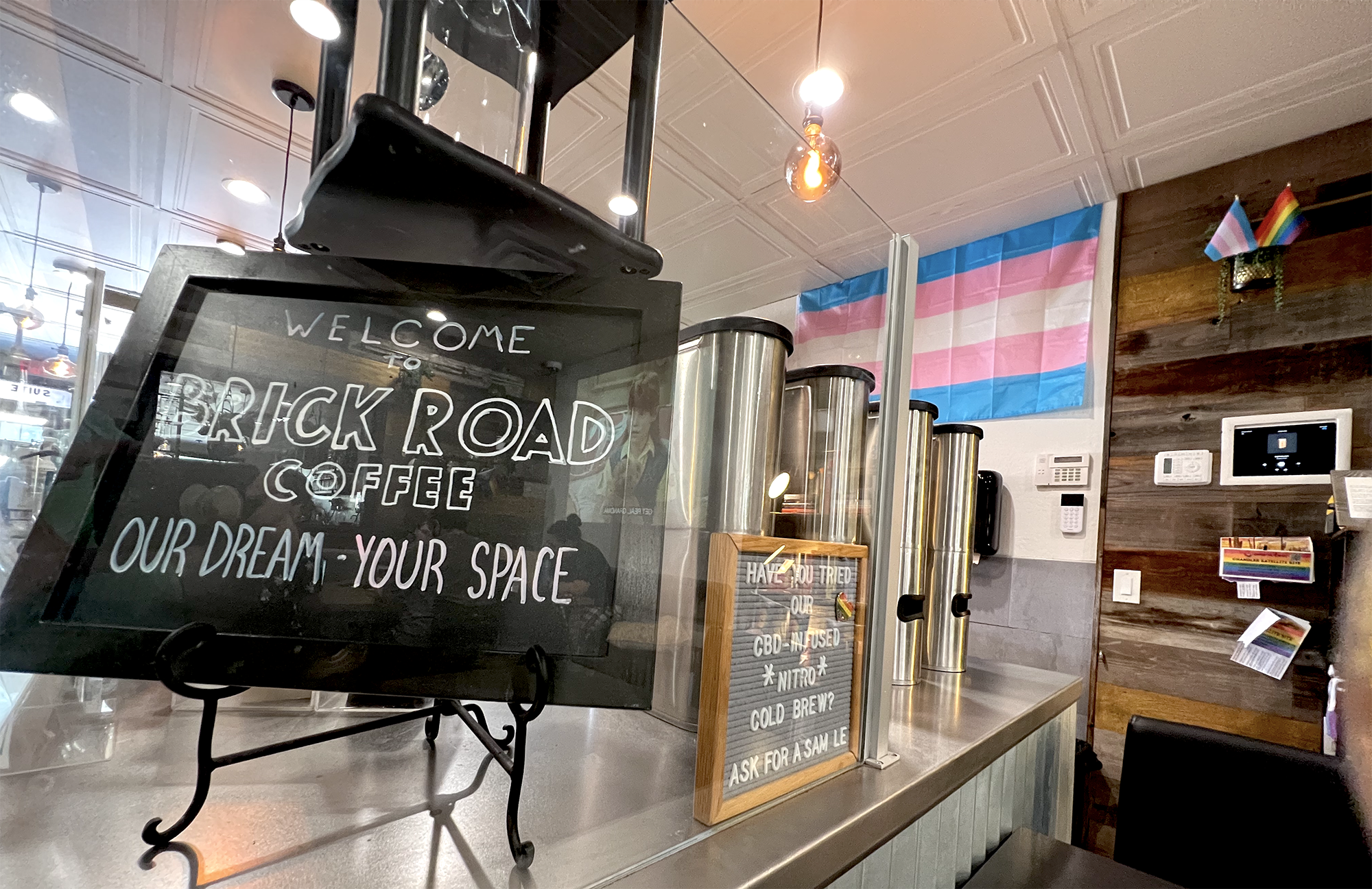 a small, black chalk sign reads 'Welcome to Brick Road Coffee, Our Dream, Your Space' It’s sitting on the counter of the coffee shop A blue, pink, and white trans flag is on the white wall behind the counter. A trans and pride flag can also be seen on a wooden shelf next to the larger flag. Some coffee making devices can be seen on the counter. Another sign reads have you tried our CBD infused nitro brew? Ask for a sample In the glass reflection on the counter, you can make out some people sitting around the shop as Golden Girls are projected to a wall behind the counter.